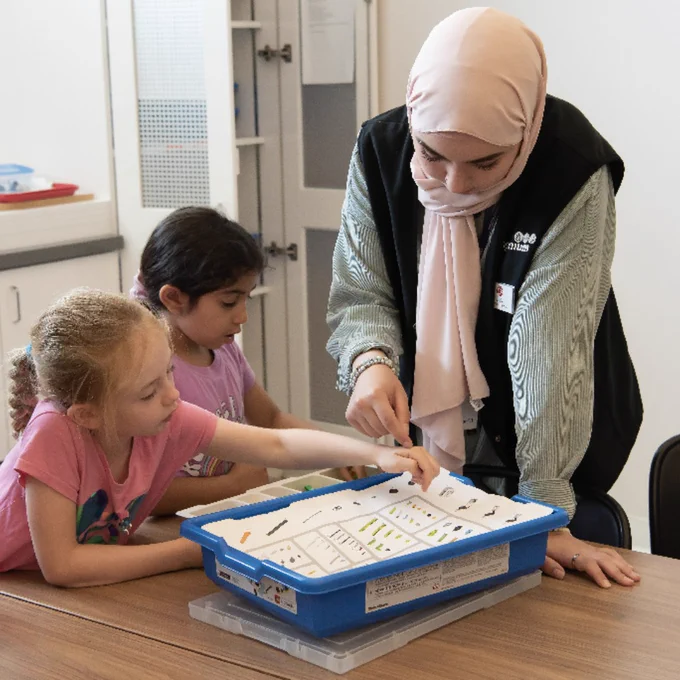 Une femme portant un foulard aide deux petites filles à découvrir le matériel pédagogique disposé dans un plateau bleu posé sur une table en chêne.