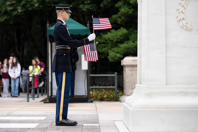 A soldier in a formal dress blue uniform stands in profile, holding two American flags at the Tomb of the Unknown Soldier. This was part of Flags In, where nearly 1,500 soldiers placed approximately 250,000 U.S. flags at every headstone and along every Columbarium Court and Niche Wall row at Arlington National Cemetery, Arlington, Virginia, Thursday, May 21, 2026.