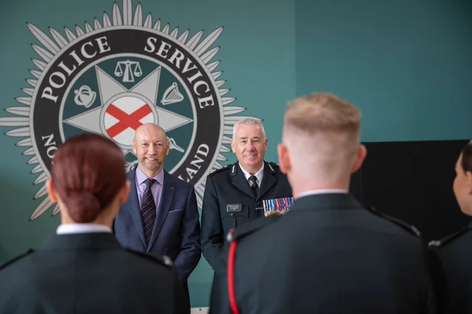 Chief Constable Jon Boutcher and Northern Ireland Policing Board Chair Brendan Mullan standing in front of two female police officers and one male police officer with the Police Service of Northern Ireland crest in the background.