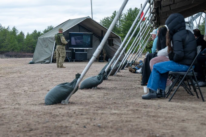 A soldier in camouflage stands near a tent with a screen, addressing seated civilians outdoors on a dirt ground.