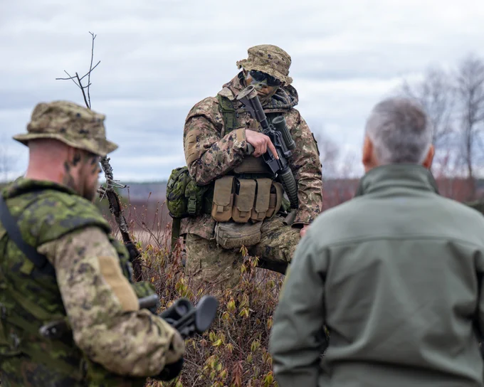 A soldier in camouflage gear holding a rifle while speaking to two people in an outdoor field.