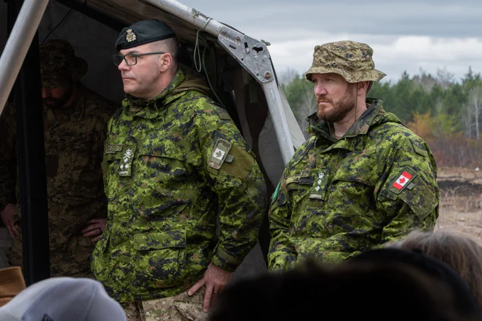 Two Canadian officers in camouflage uniforms listen to a briefing outside a tent.
