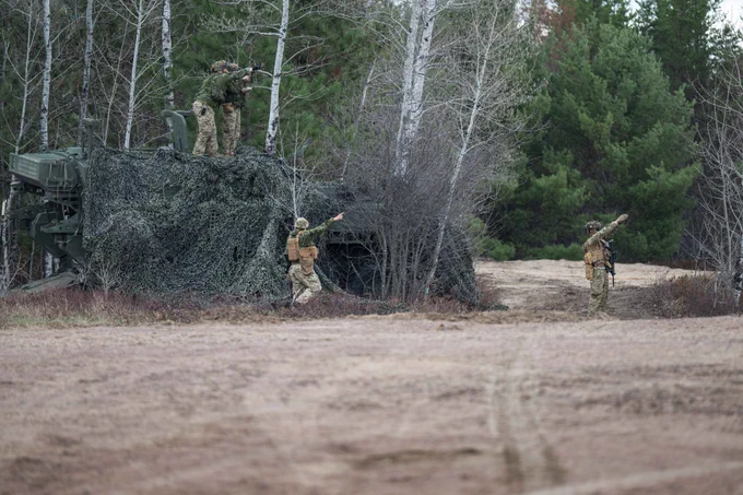 Soldiers in camouflage gear conducting a training exercise near a camouflaged military vehicle in a forest clearing.
