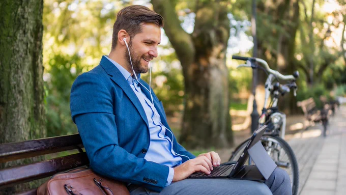 A person on a bench smiling and typing on a laptop with headphones in, with a computer bag on the bench to the left and a bike propped up in the back right.