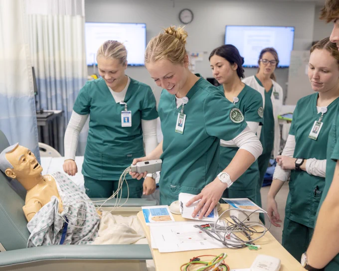 A group of medical students in green scrubs surrounds a hospital bed with a medical mannequin. They are engaged in a clinical training session, examining and discussing equipment. Monitors and medical supplies are visible in the background.