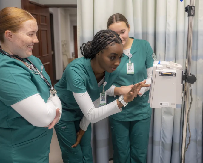 Healthcare students in green scrubs are gathered around medical equipment, engaging in discussion. They appear focused and collaborative in a clinical setting.