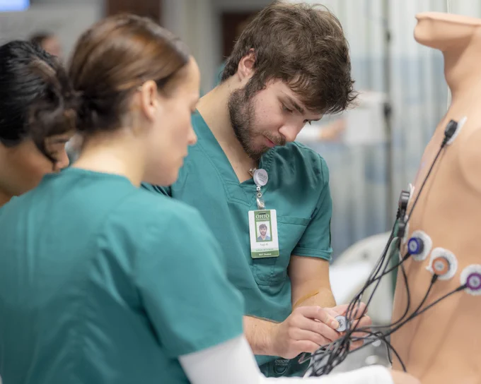 Healthcare students in green scrubs practice medical procedures on a human-like mannequin equipped with electrodes in a clinical setting.