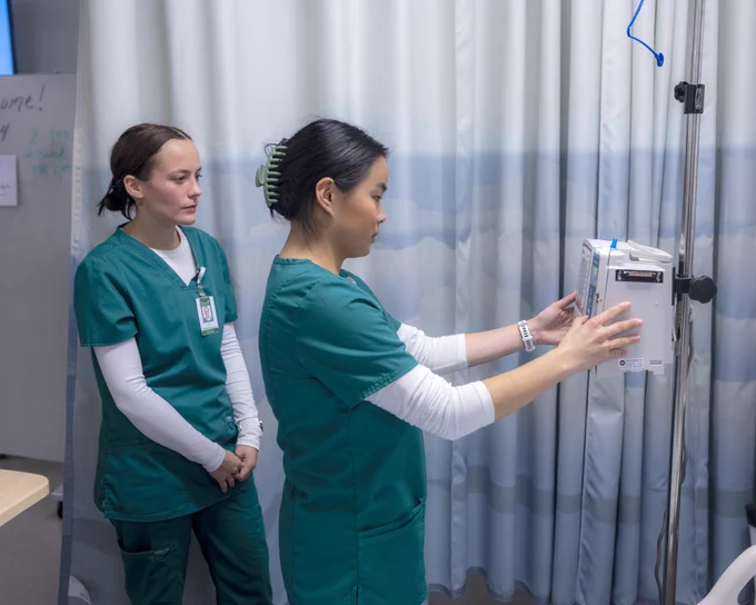Two medical students wearing green scrubs stand in a hospital room. One is adjusting an IV infusion pump, while the other observes. There is a whiteboard and curtain in the background.