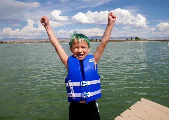 A child wearing a life jacket smiles and cheers with his hands in the air in front of a large lake on a sunny day.