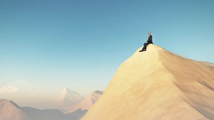 Image of a person in a suit sitting on top of a sandy hill in the desert. 