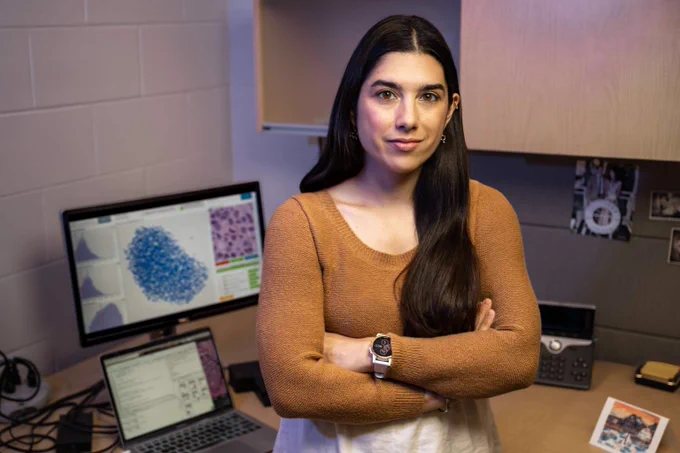 Scientist standing confidently in a lab office with arms crossed, computer screens displaying scientific data and microscopic images behind her.
