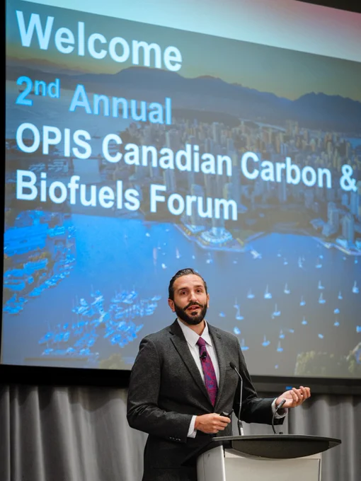 A person in formal attire stands at a podium, speaking at the 2nd Annual OPIS Canadian Carbon & Biofuels Forum. A projected background displays a cityscape and the forum's title.