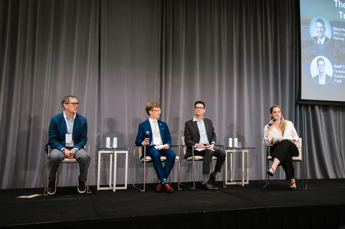 A panel of four people seated on a stage with gray curtains in the background. Each person is wearing business attire, and one of them holds a microphone. There's a display in the top right corner showing text and small images. Small tables are placed beside each chair.