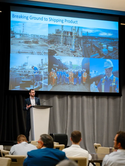 A person stands at a podium presenting to an audience. Behind them is a large screen displaying a presentation titled "Breaking Ground to Shipping Product." The screen shows various images of an industrial facility, workers in safety gear, and a container with "Dow" visible. Audience members are seated, facing the speaker.