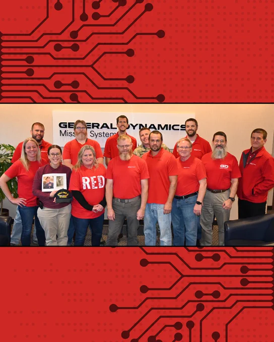 A group of people in red shirts are standing together, posing for a photo in front of a "General Dynamics Mission Systems" sign. They appear to be in an office setting. One individual is holding a framed image.

GDMissionSystems.com/Military 