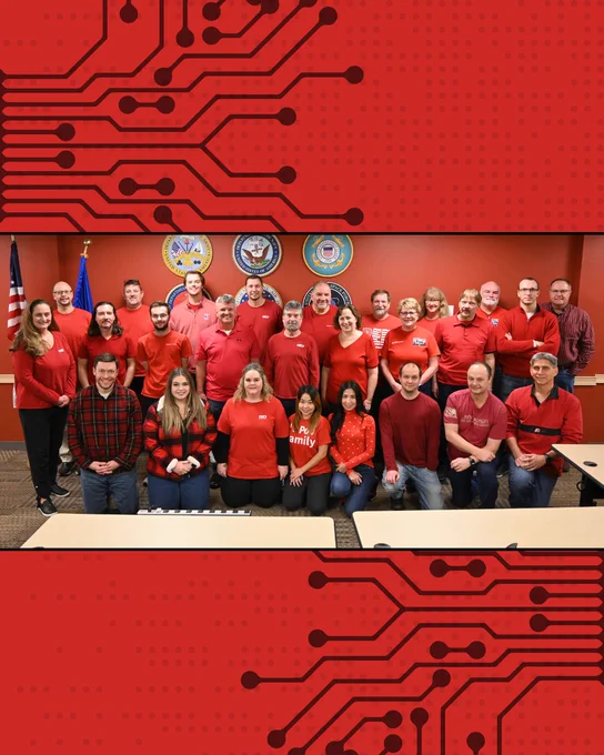 A group of people wearing red shirts is standing and sitting in a room with an American flag and official seals on the wall. The background features a red and black circuit board design.

GDMissionSystems.com/Military 