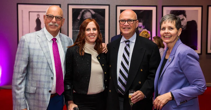 Four people are standing together, smiling, in a warmly lit room with framed photographs on the walls. They are dressed in formal attire.