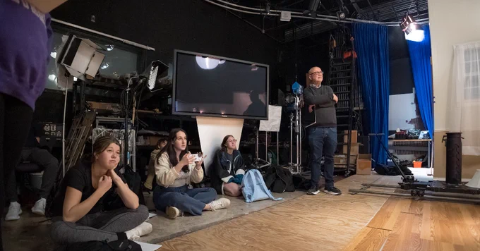 A group of people is seated on the floor in a studio setting, focused on watching a presentation or activity. Behind them, various equipment, including a large monitor and tripods, is visible. A person stands nearby, observing with arms crossed. The area is dimly lit with a few bright lights and blue curtains in the background.