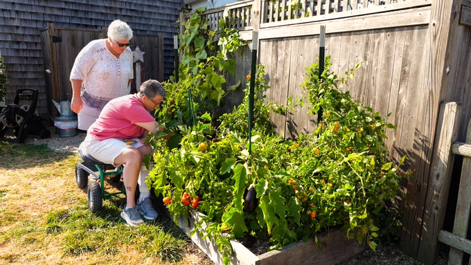 Patty Ericson-Taylor and Dawn Ericson-Taylor in their outdoor garden. Patty is using a seated device with wheels to assist with her gardening in the raised bed.