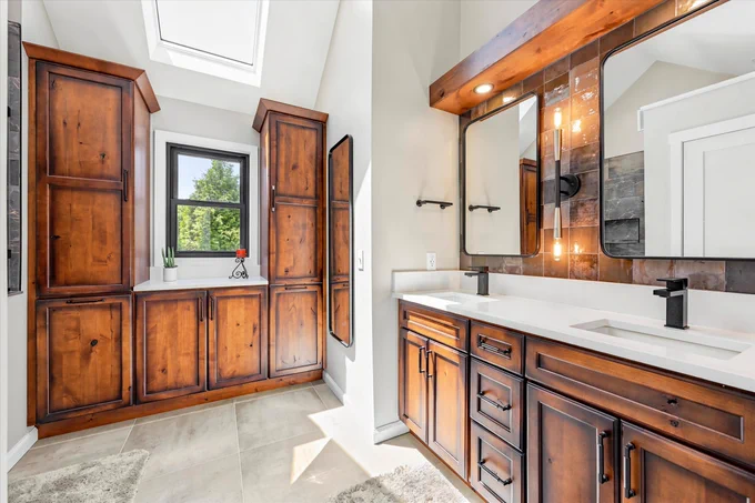 A bright and modern bathroom featuring wooden cabinets, dual sinks, and a skylight above. The space is well-lit with natural light and has a minimalistic and elegant design.