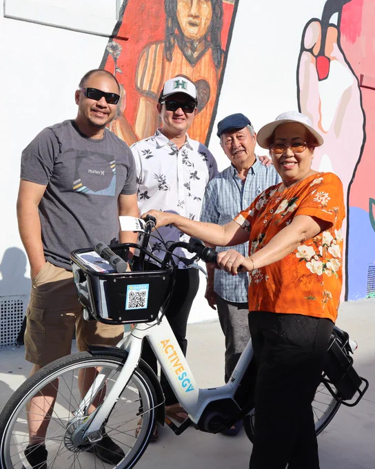 A group of four people standing by a mural with vibrant colors. One person is holding a bicycle labeled "Active SGV." Some individuals are wearing hats and sunglasses.