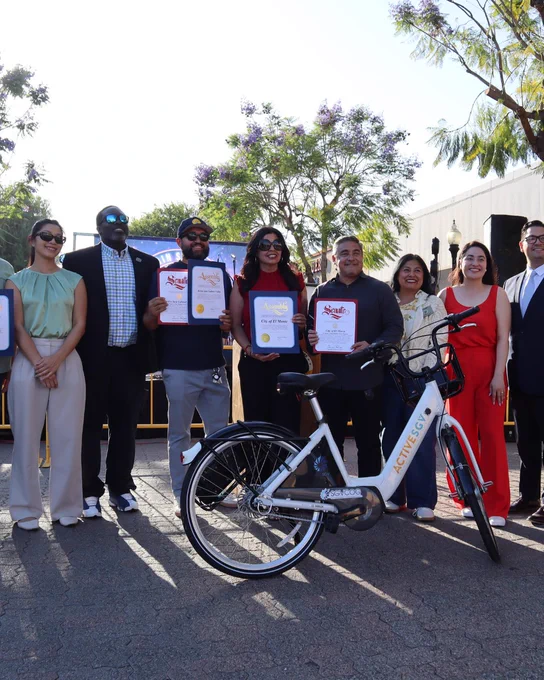 A group of people stand together outdoors, smiling and holding certificates. In front of them is an Active SGV bike. Trees and a building are visible in the background.