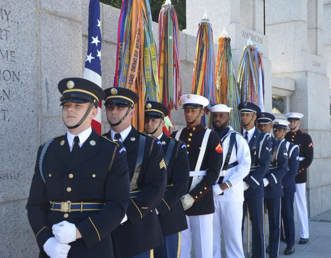 A United States military color guard of young men in the uniform of every branch holding flags in a line in an outdoor military memorial.