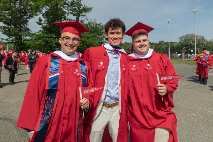 Three graduates in red robes and white hoods pose together outdoors; one wears a Marine Corps stole and others hold red pennants.