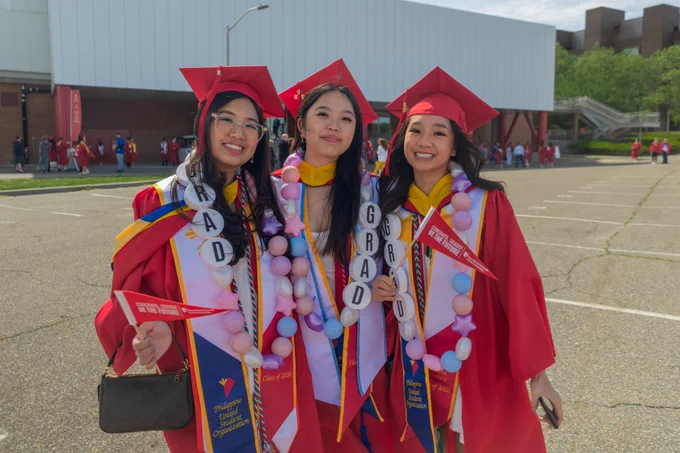 Three graduates in red caps and gowns smile together outdoors, wearing custom candy leis and holding small red pennants.