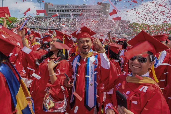 Graduates in red gowns cheer excitedly at a Stony Brook University stadium ceremony as red and white confetti rains down.