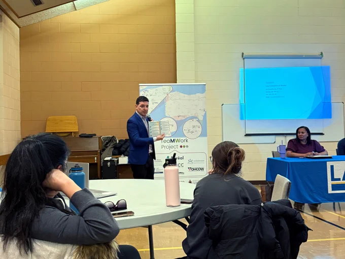 Un ponente se encuentra de pie al frente de una sala comunitaria, sosteniendo un documento mientras habla a un pequeño grupo de trabajadores migrantes sentados en las mesas. Detrás del ponente se ven una pancarta del «Proyecto TeaMWork» y una diapositiva proyectada, mientras los asistentes escuchan.