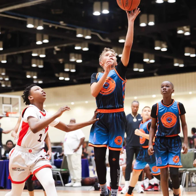 A youth basketball game at the AAU Basketball Memorial Day Classic, being held at the Orange County Convention Center, where a player in a blue uniform is making a layup. Another player in a white uniform is reaching to block the shot. Spectators and players are visible in the background on the court.