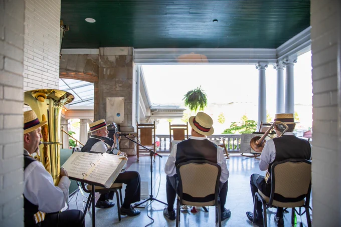 A small band is performing on a covered porch with a scenic view. The musicians are wearing straw boater hats and vests, playing brass instruments. Music stands and a microphone are set up in front of them. The porch features columns and overlooks a landscape with greenery.