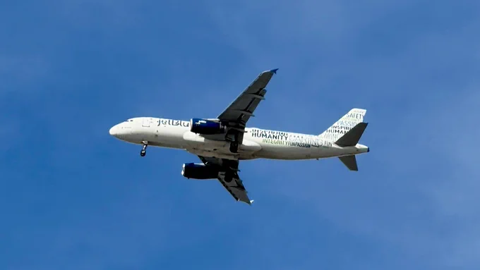 An airplane in flight against a blue sky, displaying the JetBlue logo and branding.