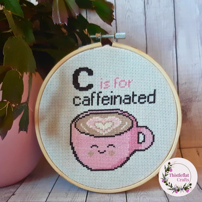 Funny cross stitch in wooden hoop reading 'C is for caffeinated' featuring cute pink smiling coffee cup, styled with a plant on rustic wooden background – handmade snarky embroidery wall art