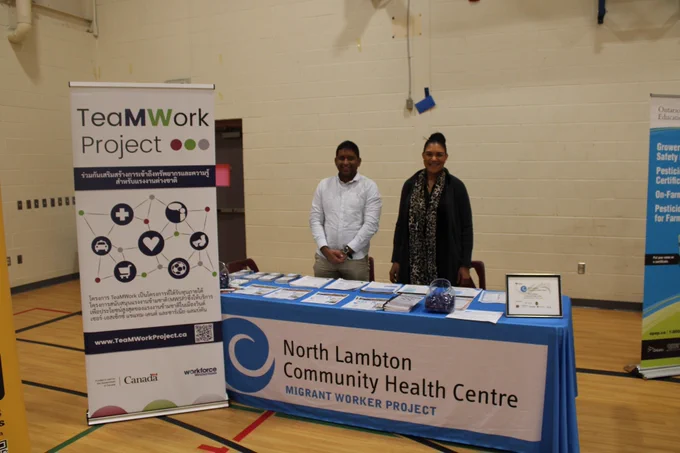 Two service providers stand behind an information table displaying pamphlets and resources at a community event. A “TeaMWork Project” banner and a “North Lambton Community Health Centre – Migrant Worker Project” tablecloth are visible, highlighting services available to support migrant workers.

