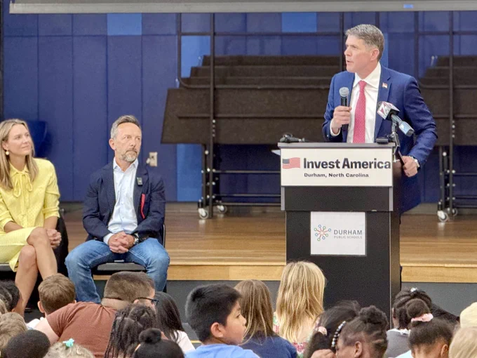 Treasurer Briner stands at a podium with an "Invest America" sign, speaking into a microphone, while another person sits nearby. The event takes place in Durham, North Carolina, with an audience of people seated in the foreground.