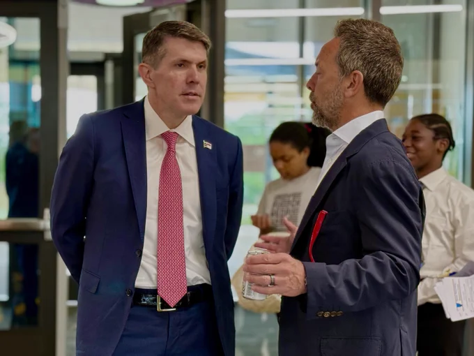 Treasurer Briner and Brad Gerstner are engaged in conversation in an indoor setting. One is wearing a blue suit and red tie, while the other is in a dark blazer holding a bottle. In the background, two other people are present, one looking at a phone and another smiling.