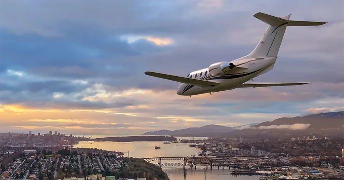 A business jet flies over a scenic cityscape at sunset. Below, there are urban areas, bridges, and open water, with distant mountains under a partly cloudy sky.