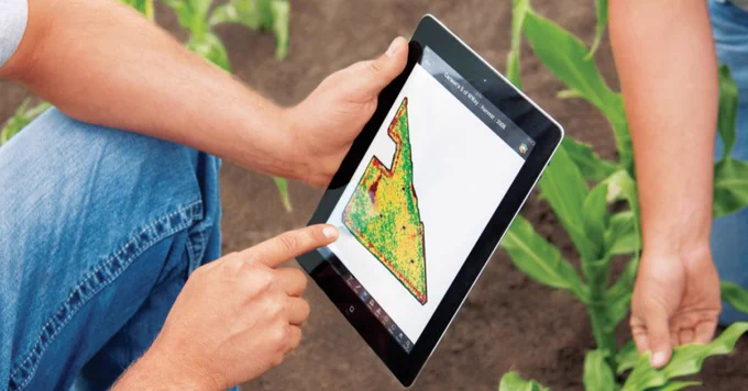 A person is using a tablet outdoors, displaying a colored map or diagram, while another person is tending to green plants in the soil nearby.