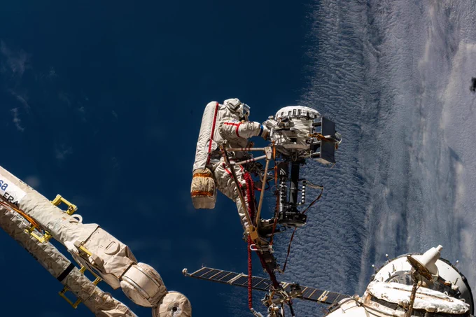 A photo of Roscosmos cosmonaut Sergey Ryzhikov on a spacewalk outside of the International Space Station. His spacesuit and the parts of the station in the scene are off-white in color, contrasting with the cool water and clouds of Earth in the background. Credit: NASA