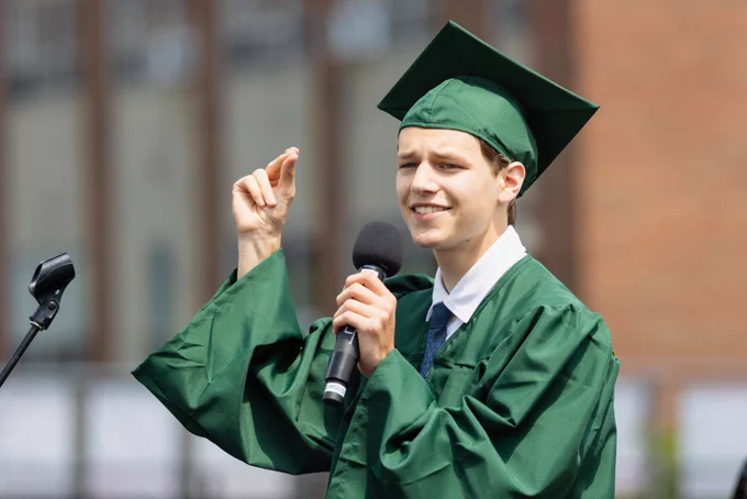 A young graduate in a green cap and gown delivers a speech outdoors. He holds a microphone and gestures confidently, adding a dynamic element to his presentation.