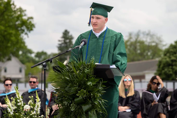 A graduate in a green cap and gown speaks at a podium during an outdoor ceremony, surrounded by leafy decorations. The audience in robes listens attentively on a sunny day.