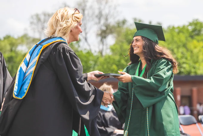 A proud graduate receives her diploma with a smile during an outdoor ceremony, sharing a handshake with a faculty member in regalia. The atmosphere is celebratory and joyful.