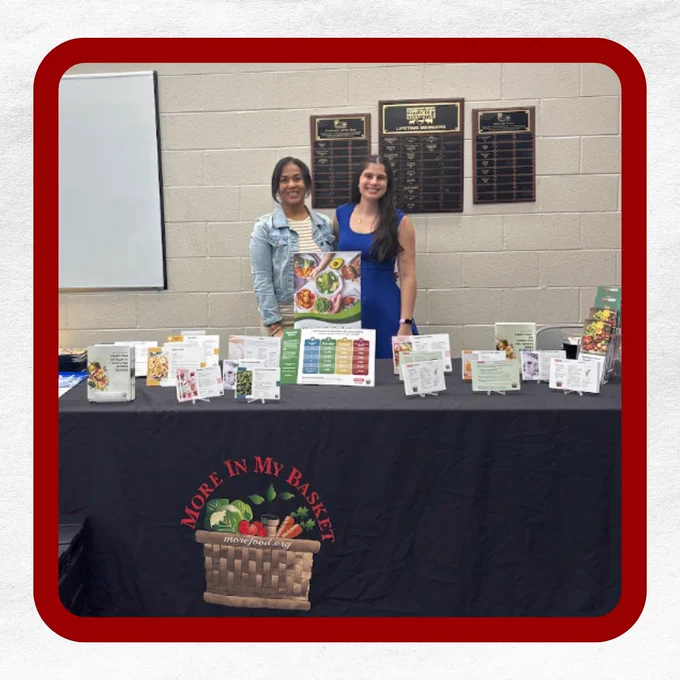 Two women stand behind a display table with food and nutrition brochures, with "More In My Basket" on the tablecloth.