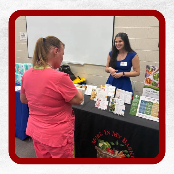 Two women interacting at a table with informational brochures and a "More In My Basket" logo.