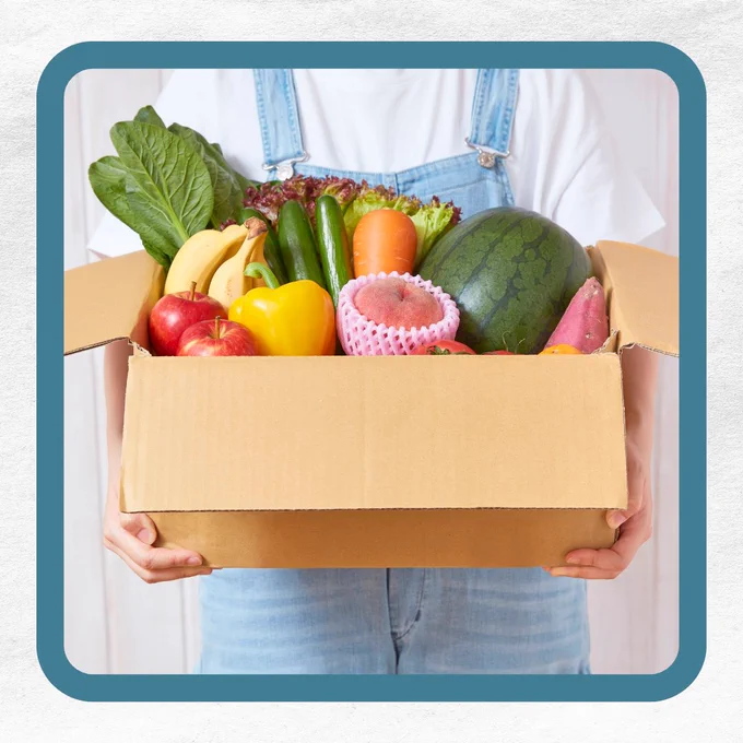A person holding a cardboard box filled with assorted fruits and vegetables, including a watermelon, bananas, apples, and a yellow bell pepper.