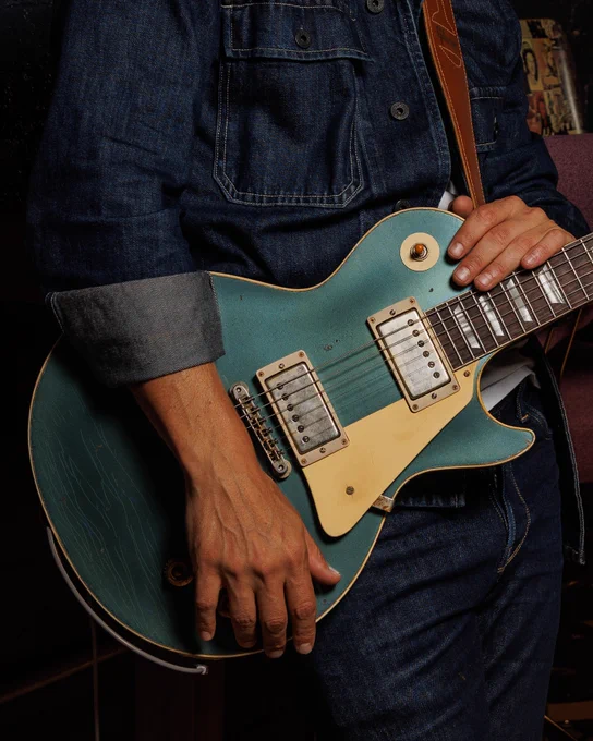 A person in denim clothing holds a blue electric Gibson Custom '57 Les Paul Standard guitar, in a Pelham Blue Over Goldtop finish.