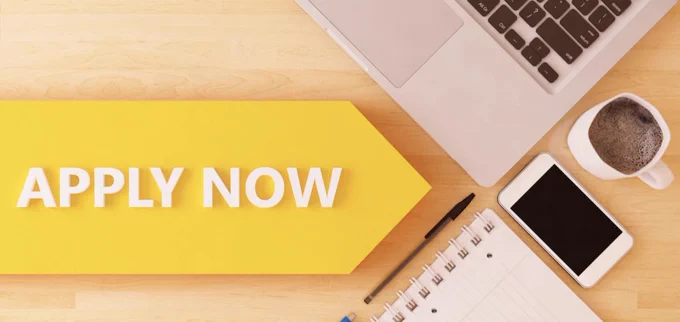 Desk with laptop, notebook, phone and cup of coffee. Text: Apply Now.