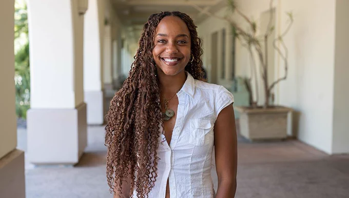 The image shows a person smiling, with long curly hair, wearing a white sleeveless shirt, standing in a shaded outdoor corridor with arches.